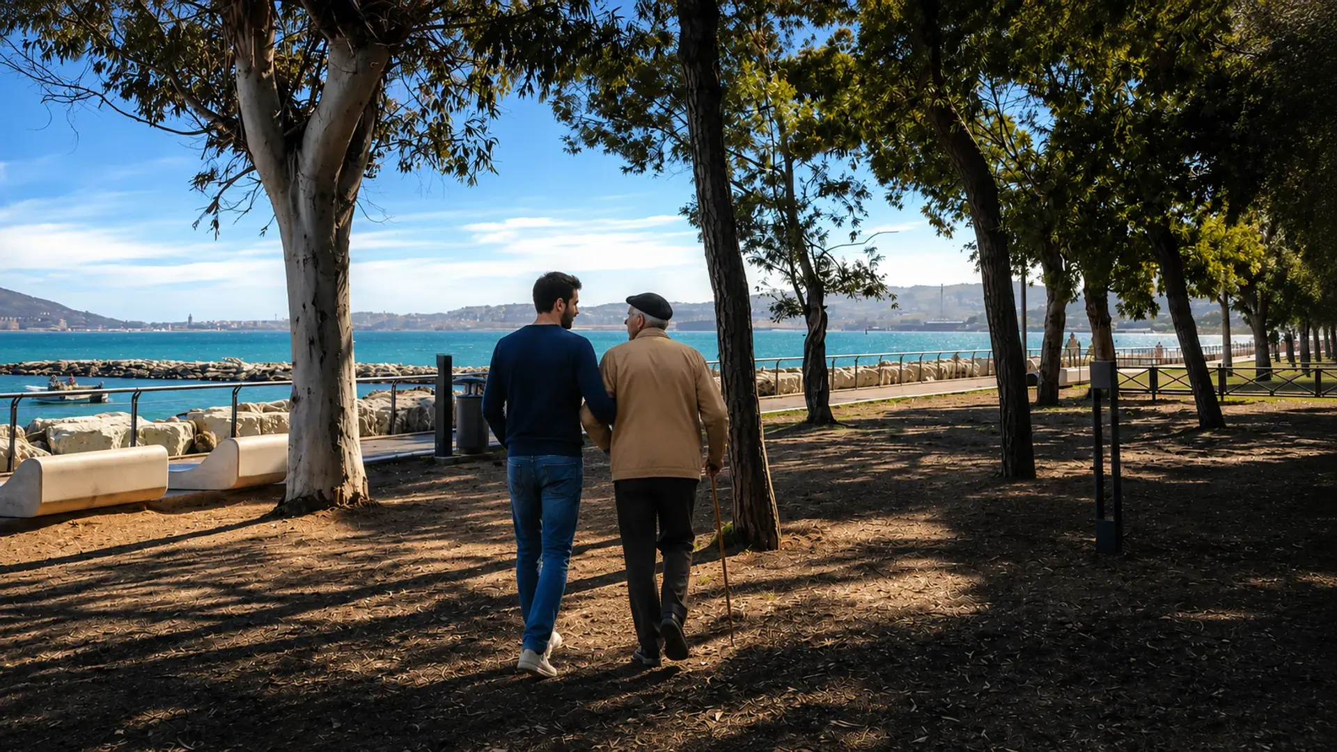 Una persona di fiducia accompagna un anziano lungo il lungomare alberato di Formia, con il golfo sullo sfondo.